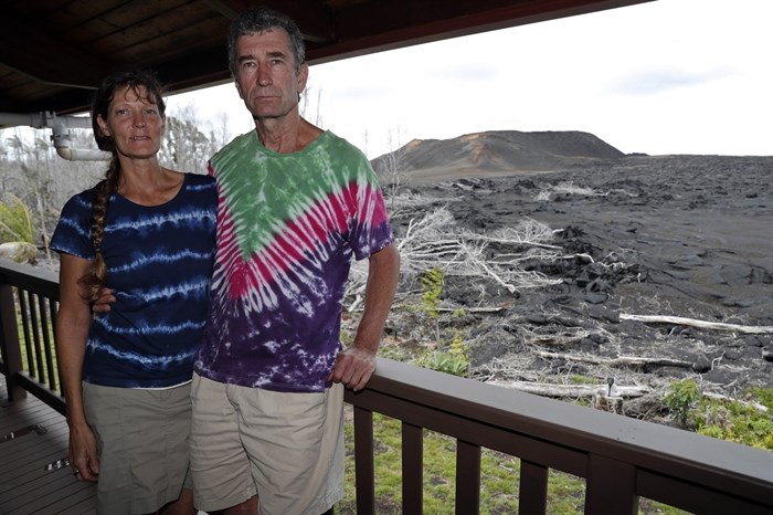 In this Tuesday, April 23, 2019 photo, with a now-dormant towering cinder cone looming in the background, Mark and Jennifer Bishop stand on the deck of their home near Pahoa, Hawaii, which was spared by the lava. The epicenter of the 2018 eruption - one of more than 20 places where the ground split open and released massive explosions of molten rock - is now in their front yard. The red-hot fluid oozed onto their property and stopped about 20 feet (6 meters) from the home. Theirs is now the last house on the street.