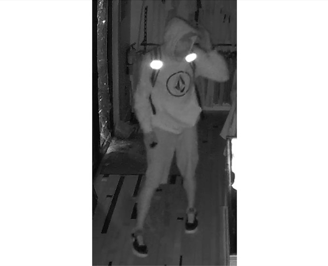 A night vision security image of a suspect with a sweater, shorts, and backpack. The sweater as a logo inside a circle.